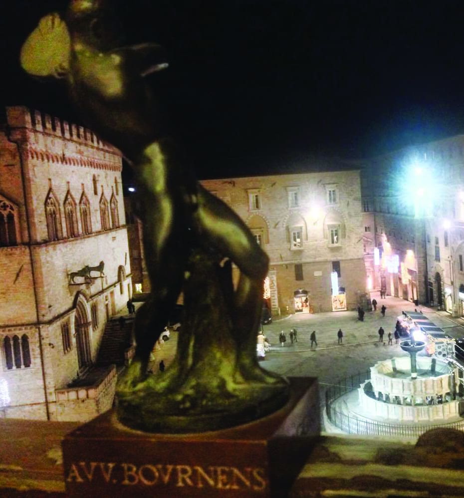 Una scultura in bronzo affacciata su Piazza IV Novembre di notte, con la Fontana Maggiore illuminata sullo sfondo